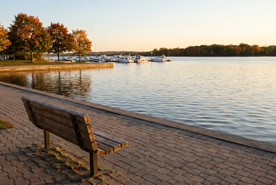 Generated scene: an empty wooden bench on a waterfront path in early evening, with a small marina and autumn maples; not a specific place.